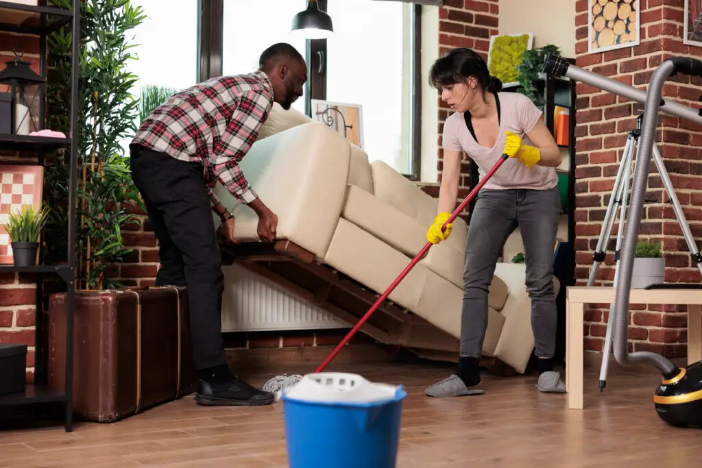 A culturally diverse couple doing bond cleaning—man lifting the sofa while the woman mops underneath it, inside a modern apartment setting in Perth.