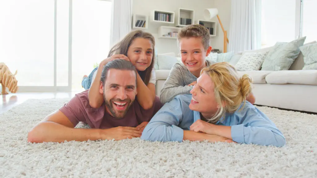 A cheerful family of four—parents, daughter, and son—lying together on a spotless white carpet in their Perth home, smiling and relaxed, illustrating the comfort and peace of mind that comes from professional carpet cleaning in Perth.
