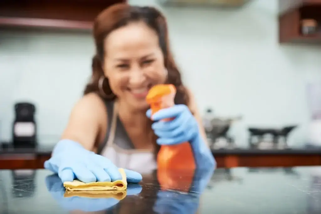 Smiling woman wearing blue gloves cleaning a kitchen in a Perth home with spray and microfiber cloth during bond cleaning.